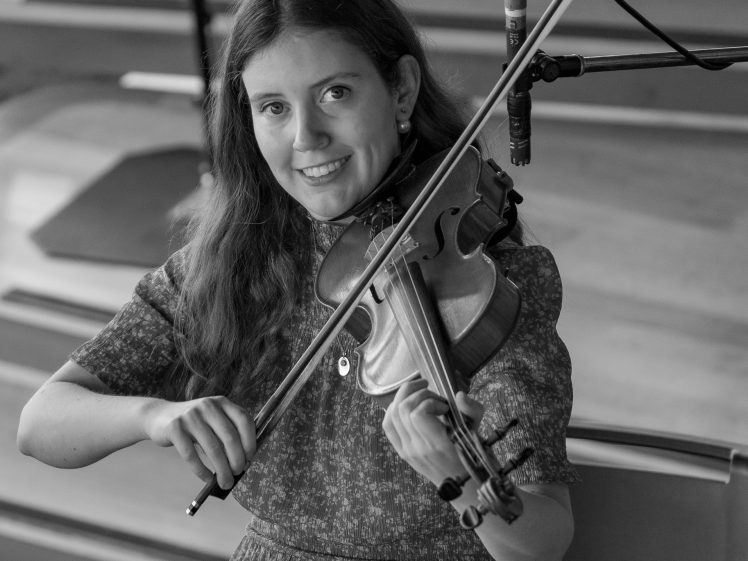 Image of A young woman with long hair plays the violin and smiles at the camera. She is standing in front of wooden steps, and a microphone is positioned next to her instrument. The image is in black and white.