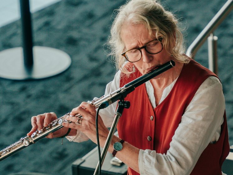 Image of An older person with wavy grey hair, glasses, and a red waistcoat plays a silver flute indoors, standing by a black case and metal railings on a dark carpeted floor.