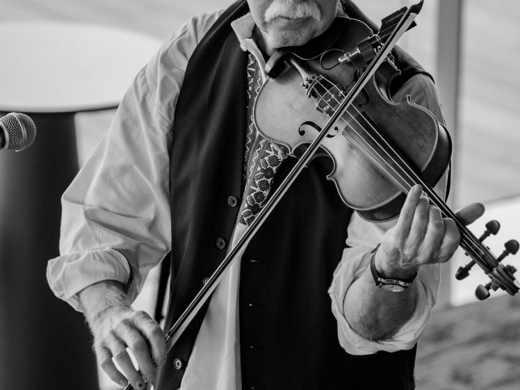 Image of A man wearing a hat and waistcoat plays the violin indoors. He has a moustache and is focused on his performance. The image is in black and white, with a blurred background and a microphone nearby.