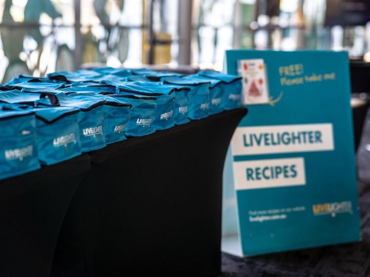 Image of A row of blue tote bags sits on a black table next to a large sign that reads LIVELIGHTER RECIPES in a bright indoor setting. The sign offers free recipe packs.