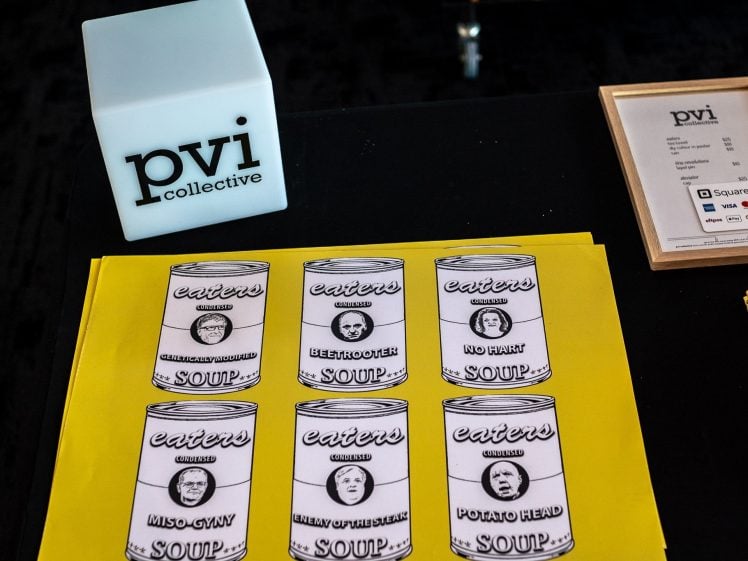 Image of A table displays a light cube with pvi collective on it, a framed sign, pens, and a yellow sheet featuring six illustrated soup tins labelled with puns like Beetroot Soup and Potato Head Soup.