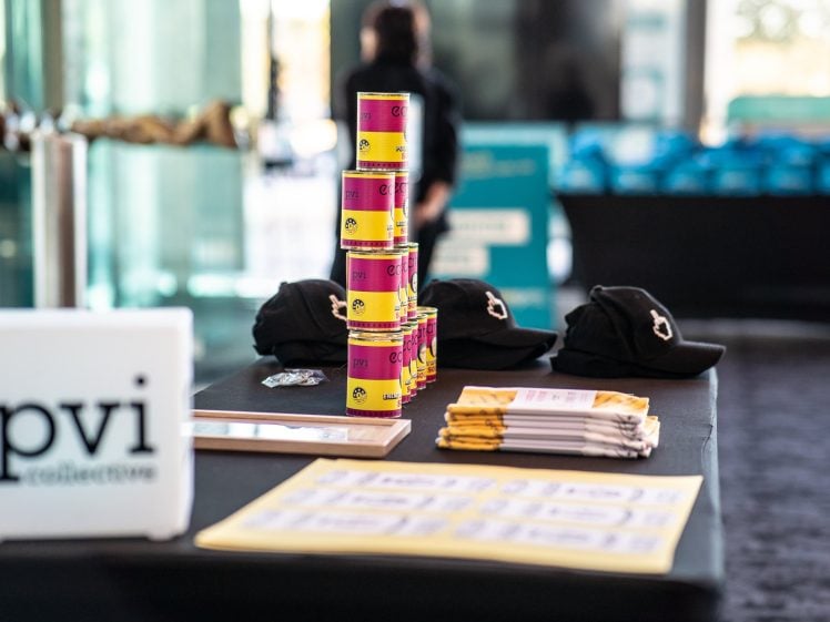 Image of A table displays stacked tins, black baseball caps, yellow booklets, and a sign reading pvi collective at an indoor event space with blurred people and items in the background.