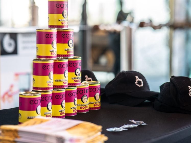 Image of A stack of colourful tinned soups labelled eat eat eat next to two black baseball caps with a white raised middle finger design, all displayed on a black table indoors.