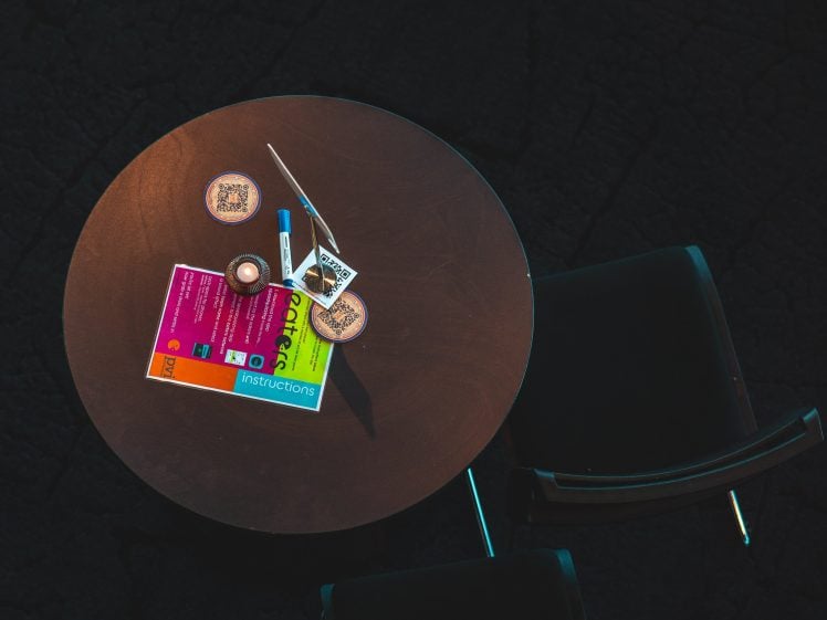 Image of A round table with two chairs holds a candle, coasters, a marker, a pen, a flyer, and some small cards, seen from above. The background is dark, emphasising the tabletop items.