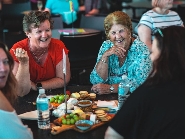 Image of Four women sit around a table, smiling and laughing together. On the table are water bottles, cheese, crackers, fruit, and dips. The group appears to be enjoying a friendly, casual gathering indoors.