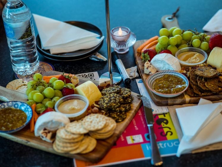 Image of Two wooden boards with assorted cheeses, biscuits, grapes, strawberries, dips, and a jar of pickle. A bottle of water, candle, serviettes, and colourful leaflets are also on the table.