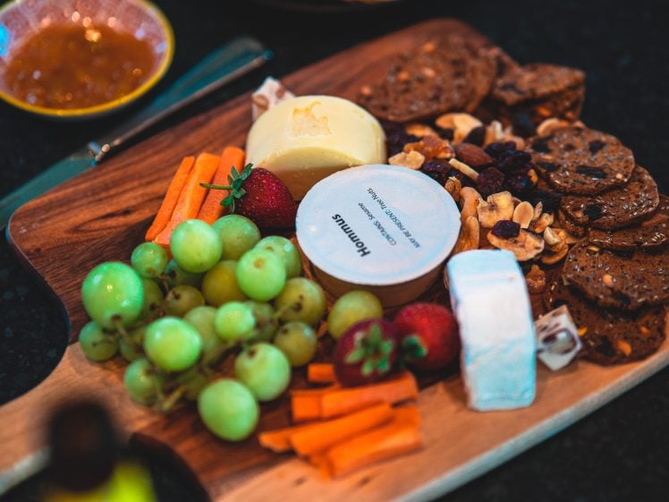 Image of A wooden board with green grapes, strawberries, cheese, carrot sticks, crackers, walnuts, dried fruit, and a round container of houmous, set on a dark surface with a knife and a small dish in the background.