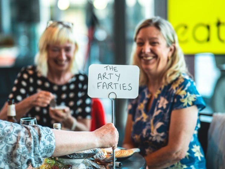 Image of Two smiling women sit at a table, one in a black polka dot shirt and the other in a floral shirt. A sign in the foreground reads THE ARTY FARTIES. The scene appears casual and friendly.
