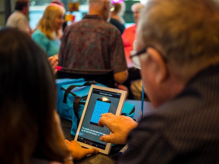 Image of A man uses a tablet, entering a PIN on the screen, whilst sitting next to a woman in a busy, brightly lit room with other people in the background.