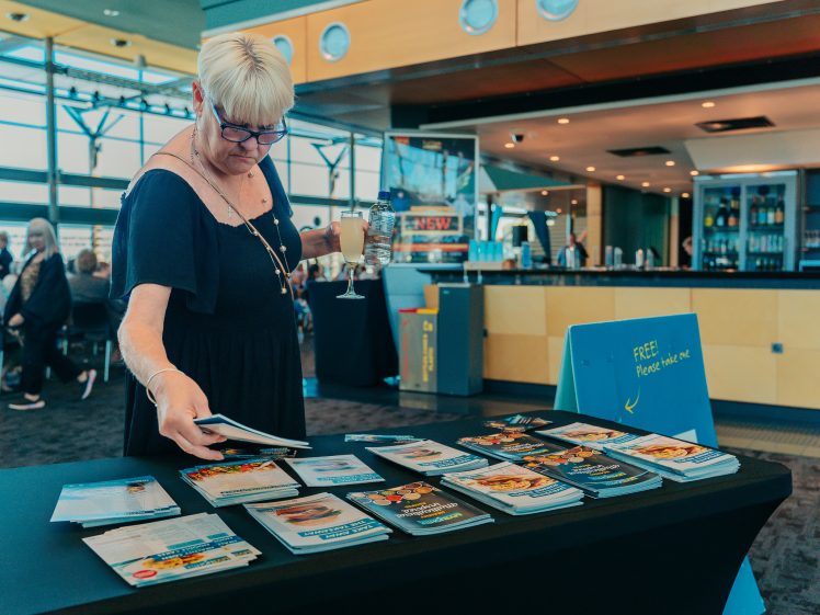 Image of A woman with short blonde hair and glasses browses brochures on a table in a modern indoor setting, holding a drink in one hand. The table sign reads FREE! and people are visible in the background.