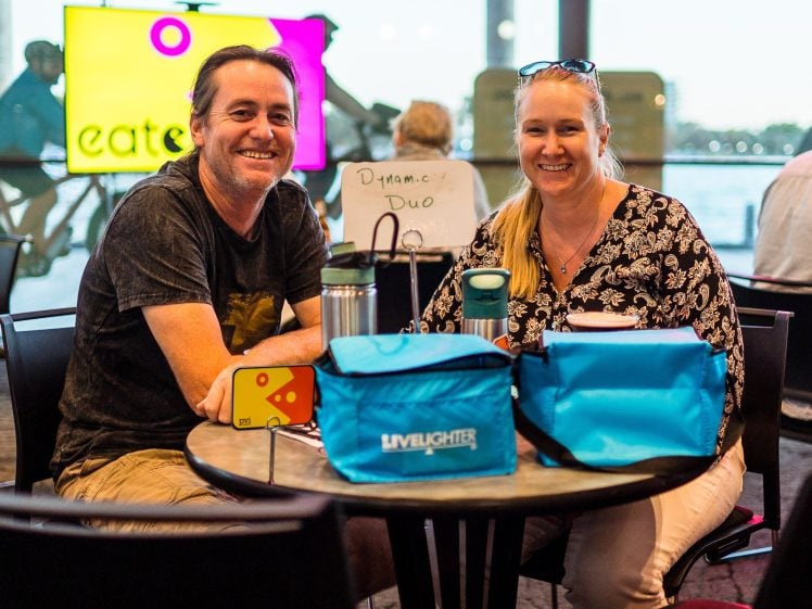 Image of A smiling man and woman sit at a table in a café with blue lunch bags and drinks. A bright eat sign and other people are visible in the background. A small sign on their table reads Dynamic Duo.