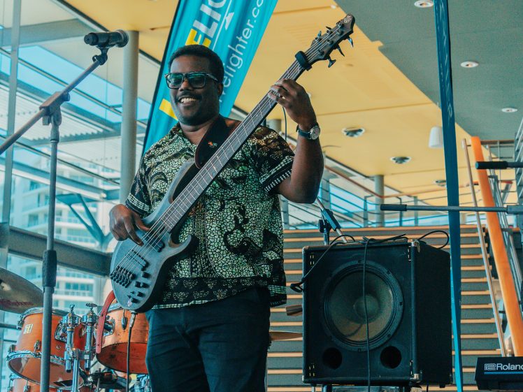 Image of A musician smiles whilst playing an electric bass guitar onstage, standing in front of a drum kit and speakers, with a blue promotional banner, microphone, and staircase in the background.