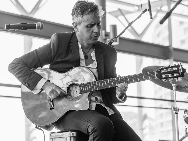 Image of A person in a suit jacket plays an acoustic guitar on stage, sitting next to a microphone and a drum kit, with large windows and buildings visible in the background. The image is in black and white.