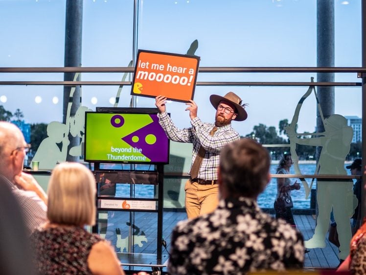 Image of A man in a cowboy hat holds up a sign that says let me hear a mooooo! while speaking to an audience seated indoors by large windows overlooking water.