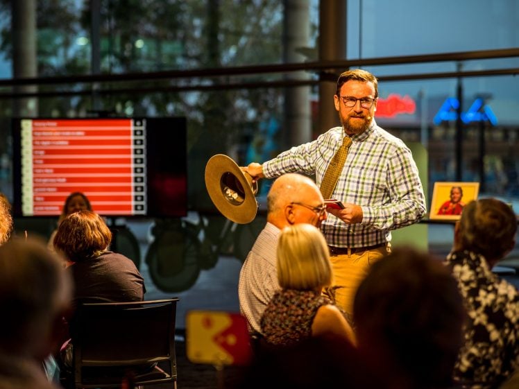 Image of A man in glasses and a checked shirt stands holding a cymbal and a card, addressing a seated audience in a brightly lit room. A large screen with red text is visible in the background.