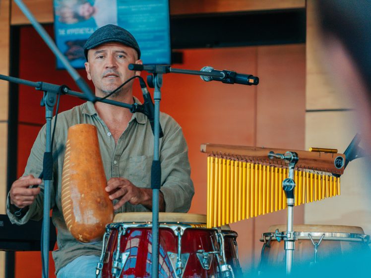 Image of A musician wearing a flat cap plays percussion instruments, including a guiro, congas, and chimes, while sitting behind microphones during a live indoor performance.