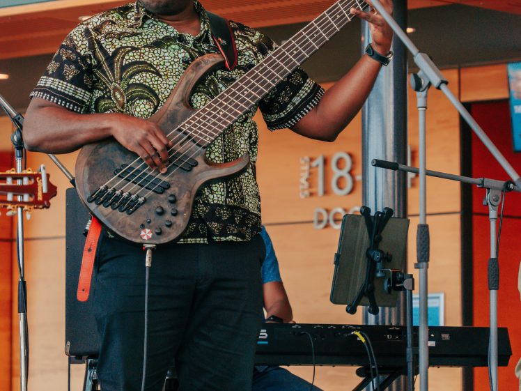 Image of A musician wearing sunglasses and a patterned shirt plays an electric bass guitar on stage, with a keyboard player behind him and microphones set up around them.