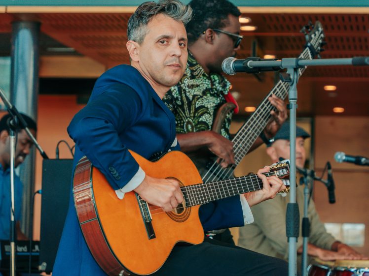 Image of A man in a blue suit plays an acoustic guitar on stage with a band. Other musicians are visible in the background, including a bassist and a percussionist, performing indoors with microphones set up.