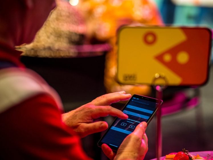 Image of A person uses a mobile phone at a brightly lit event, with a colourful sign featuring geometric shapes and a table with fruit in the background. The focus is on the person's hands and the phone screen.