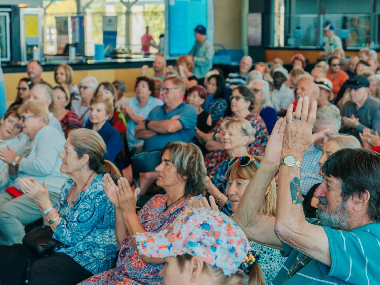 Image of A large group of older adults sits closely together, clapping and paying attention at an indoor event. The audience appears engaged, with many people wearing casual, colourful clothing.