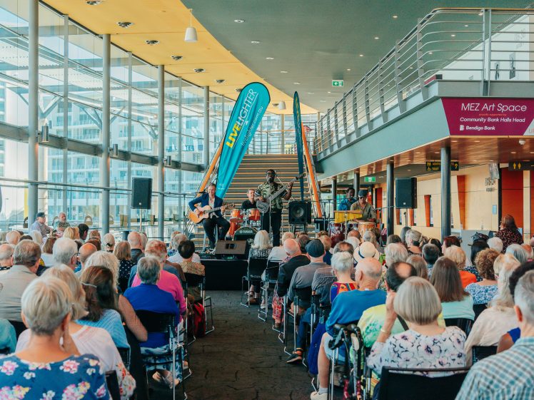 Image of A large audience sits indoors facing a small stage where musicians are performing. The venue is modern with glass walls, and a sign reads MEZ Art Space. The atmosphere is lively and attentive.