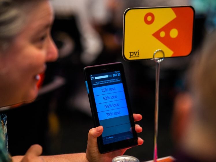 Image of A person holds a tablet displaying a quiz with options for percentage loss whilst sitting at a table. A colourful sign with geometric shapes and the letters pvi is visible on a stand nearby.