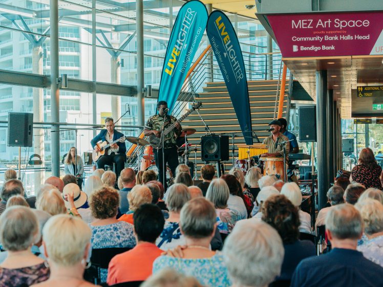 Image of A three-piece band performs on a stage in front of an audience of mostly older adults at an indoor venue with large windows. Banners and signs for the MEZ Art Space and Bendigo Bank are visible.