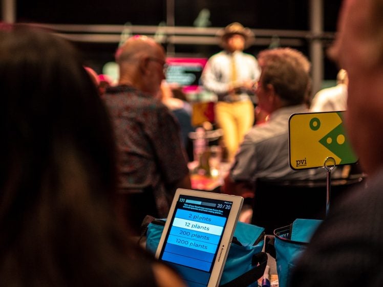Image of A group of people sit at tables, focusing on a quiz displayed on a tablet. The tablet screen shows multiple-choice answers about the number of plants. A person stands speaking at the front of the room.