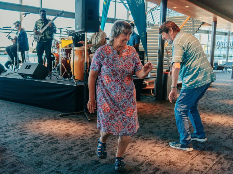 Image of A woman in a patterned dress and a man in casual clothes dance together in front of a live band performing on a small indoor stage. The setting appears to be a modern building with large windows and a staircase.