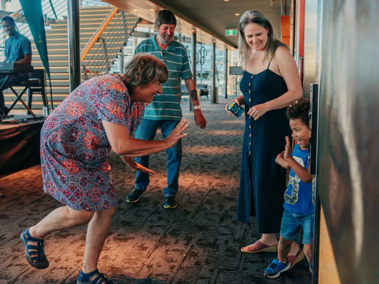 Image of A woman playfully interacts with a smiling young boy who is hiding by a wall, while another woman and a man watch and smile in a brightly lit indoor area with carpeted floors and modern décor.
