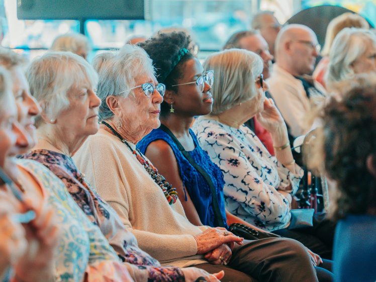 Image of A diverse group of people, mostly elderly women, sit closely together in rows, attentively watching an event. The atmosphere is calm and engaged, with soft daylight filling the room.