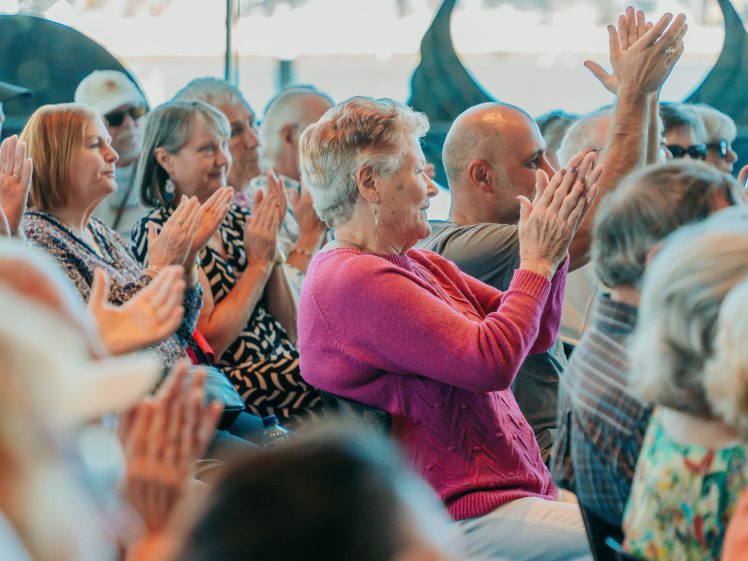 Image of A group of people of various ages sitting closely together in rows, applauding and smiling, appearing engaged and happy, as they attend an indoor event or presentation during the day.