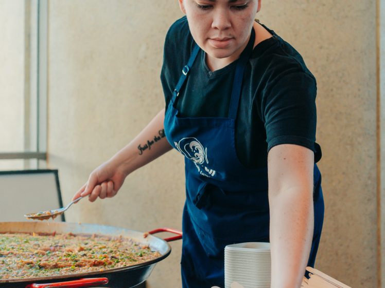 Image of A chef wearing a white hat and blue apron serves food from a large pan into a small bowl, reaching to place the bowl on a table with stacks of plates and wooden cutlery nearby.
