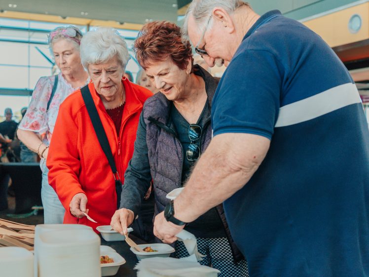 Image of Four older adults serve themselves food from a table with small dishes at an indoor event. Two women smile whilst choosing food, and a man helps serve. Serviettes and paper plates are visible in the foreground.