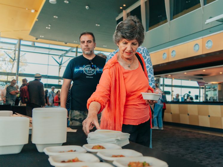 Image of A woman in an orange jacket selects food from a table with prepared dishes, whilst a man stands behind her. Other people are visible in the background inside a modern, well-lit room.