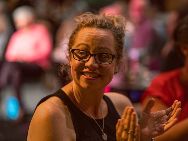 Image of A woman with glasses and curly hair pulled back smiles while clapping in a dimly lit, crowded room. She wears a sleeveless black top and a necklace. The background is blurred with people sitting and colourful lights.