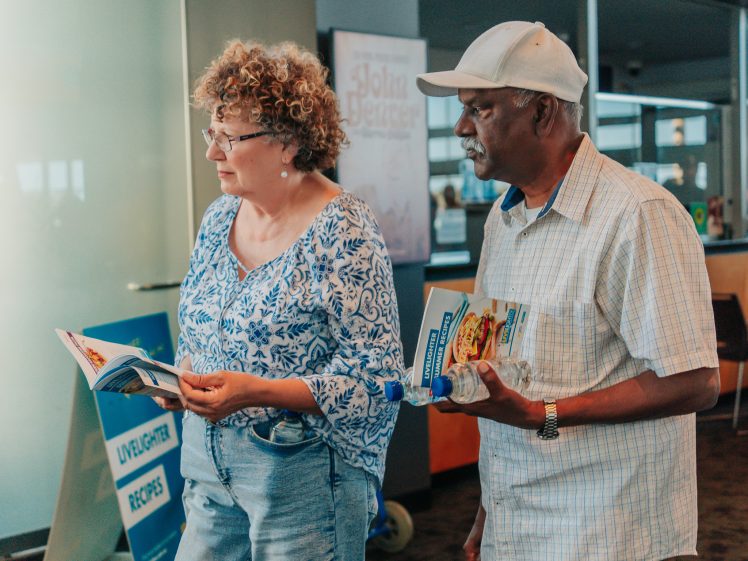 Image of Two older adults, a woman in a blue floral blouse and a man in a white cap and shirt, stand indoors holding brochures and a water bottle, appearing to read or discuss something.