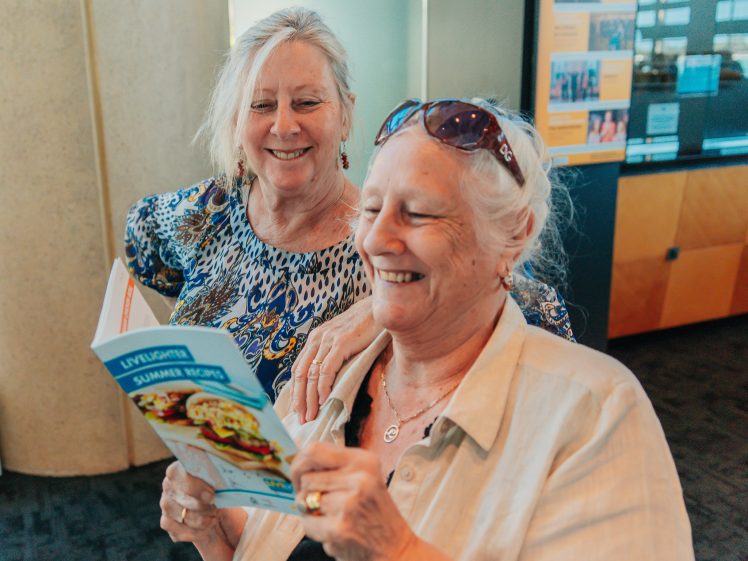 Image of Two older women smile together; one is reading a cookbook titled Lyttelton Summer Recipes while the other stands behind her, looking over her shoulder. They appear happy and engaged in a bright indoor setting.