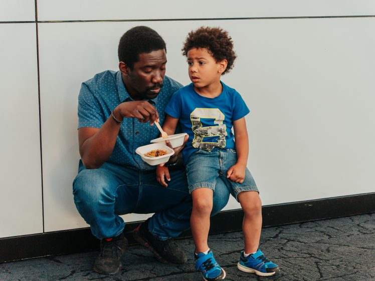 Image of A man crouches next to a young boy sitting on a ledge. The man holds a food container and is speaking to the boy, who looks slightly concerned. Both wear blue shirts and jeans. The background is a tiled white wall.