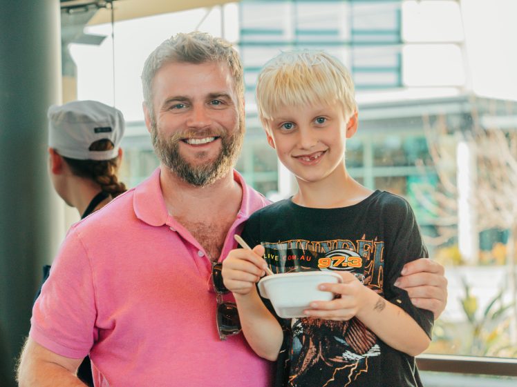 Image of A man in a pink polo shirt smiles beside a blond boy holding a bowl and spoon. They stand outdoors near a modern building, both looking at the camera. The boy is wearing a black graphic T-shirt.