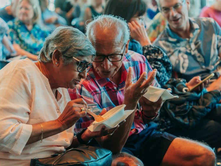 Image of Two elderly people sit closely together, smiling and sharing food from a bowl, surrounded by other seated people in a lively, social indoor setting.