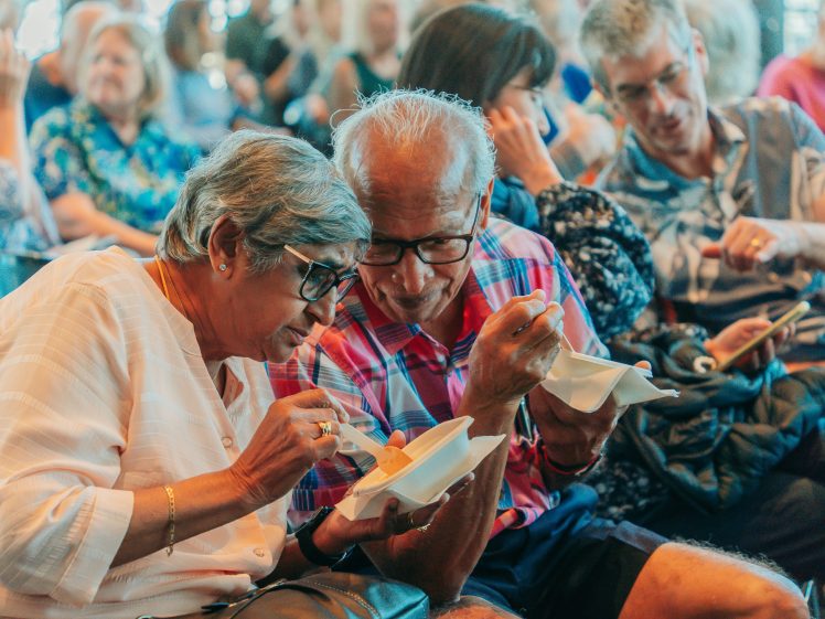 Image of An elderly couple smiles and shares a pudding at an event, sitting close together in a crowded room filled with other people who are also seated and engaged.