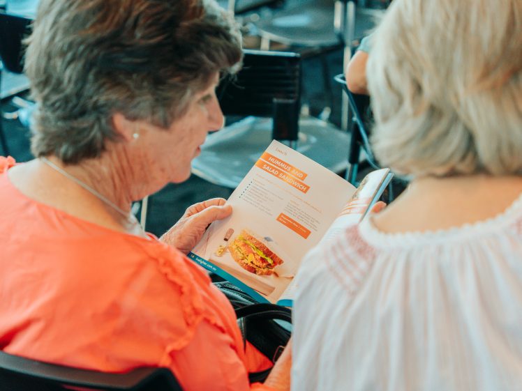 Image of Two women sit together, one wearing an orange top and the other in white, looking at a booklet featuring a recipe page with a sandwich image and a list of ingredients.