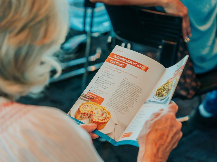Image of A person with fair hair reads a recipe booklet opened to a page featuring Courgette Quiche with Rice Crust, showing a photo of the dish and a list of ingredients and instructions.