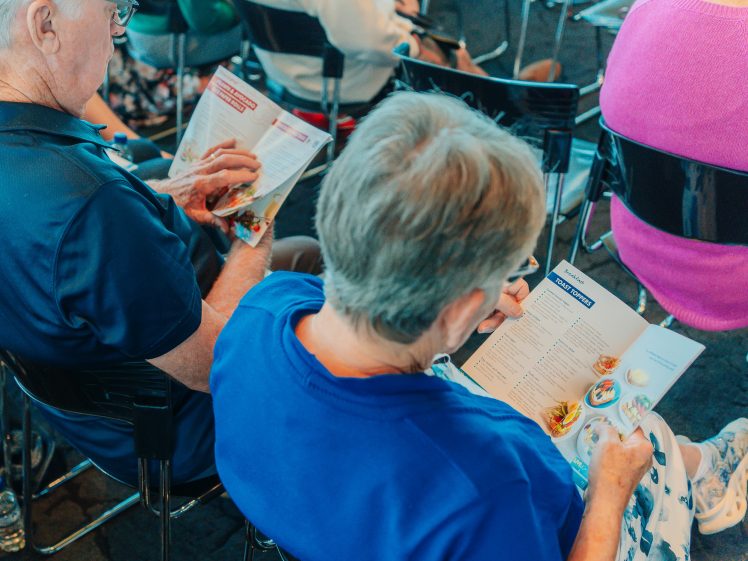 Image of Two older adults sit side by side in chairs, reading colourful brochures or menus. Other people are seated around them, and the setting appears to be a public event or seminar.