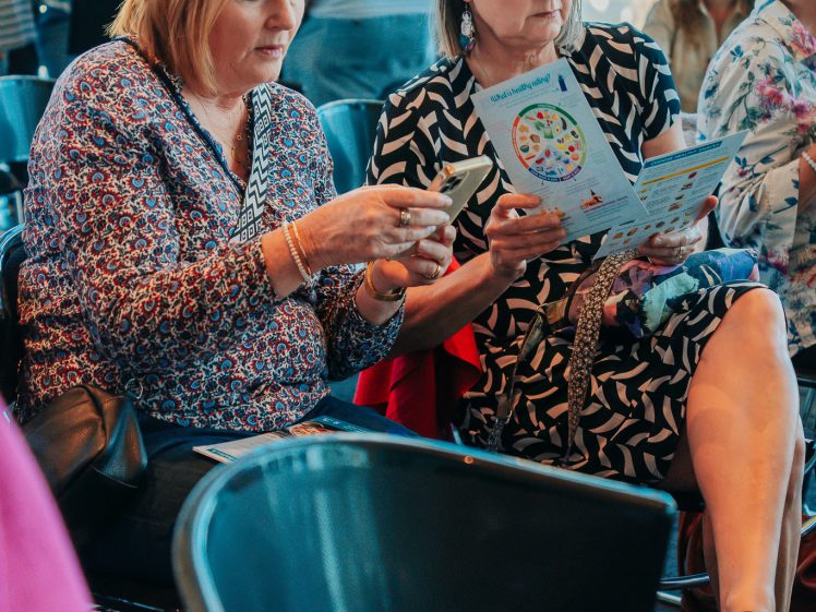 Image of Two women are sitting indoors among a crowd. One woman is looking at her mobile, while the other is reading a colourful leaflet. People and empty chairs are around them.