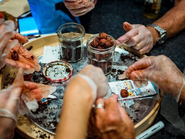 Image of Several people wearing gloves are using their hands to shape soil or seed balls and place them into glass jars on a round tray, surrounded by loose soil and tools.