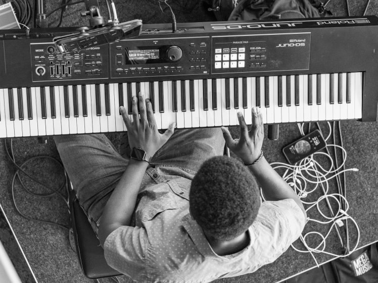 Image of A person, seen from above, plays a Roland JUNO-DS keyboard. Cables and a smartphone are on the floor nearby. The person wears a light-coloured shirt and a smartwatch. The image is in black and white.
