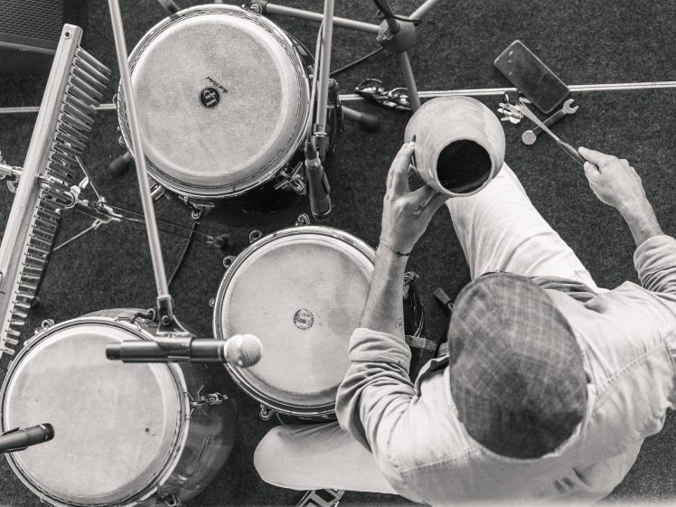 Image of Black and white top-down view of a musician playing percussion instruments, including drums, a chime bar, and a jug. The musician wears a cap and light-coloured shirt, with microphones and a mobile nearby.