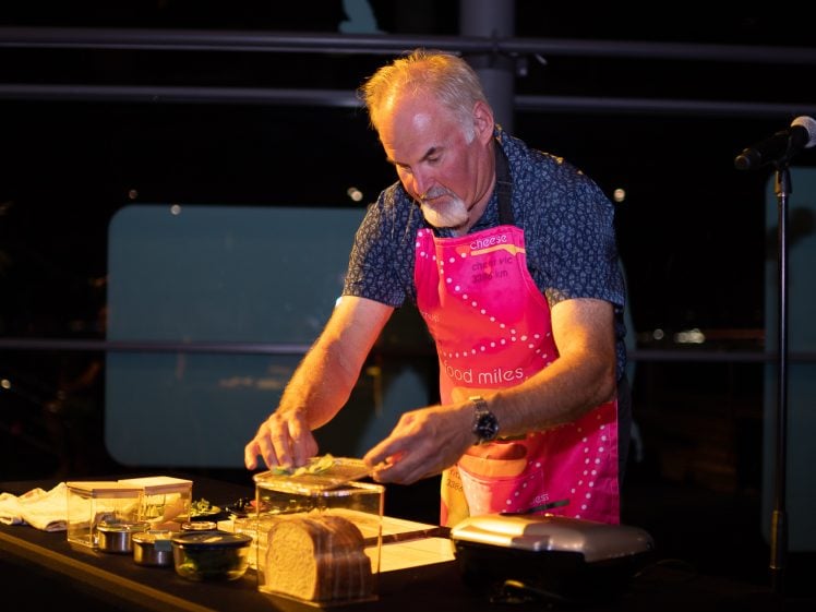 Image of A man with grey hair and a beard, wearing a pink apron, prepares food at a table with various ingredients and kitchen equipment under warm lighting.